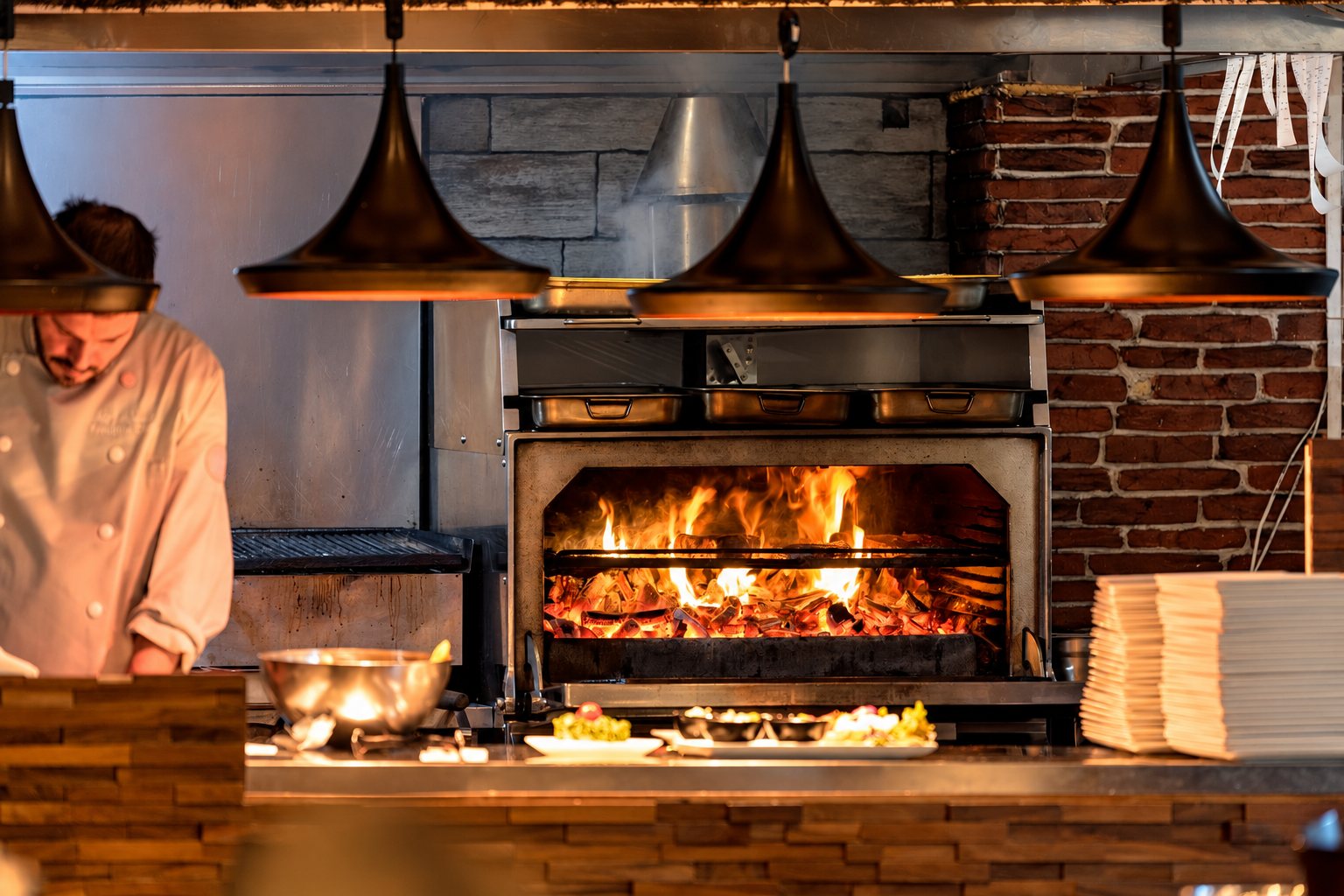 Cuisinier en tenue blanche devant un foyer à flammes dans une cuisine ouverte