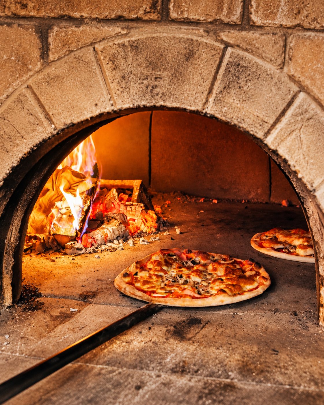 Pizzas cuisant à l’entrée voûtée d’un four à bois, flammes et braises au fond