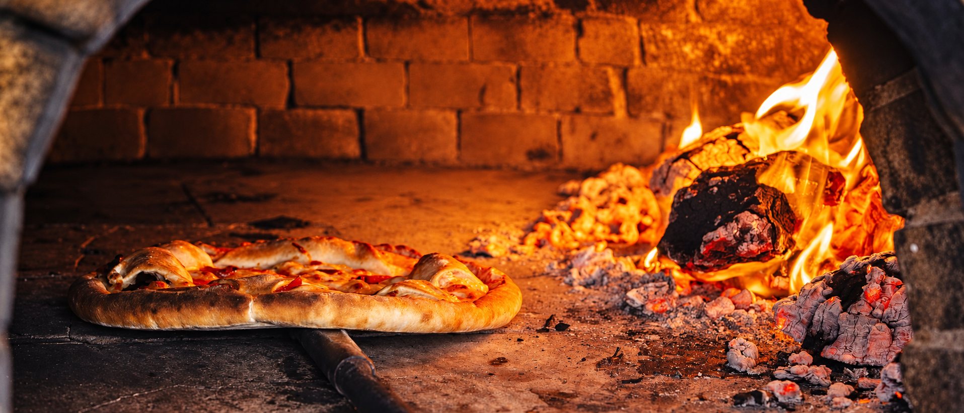 Pizza posée dans un four à bois, flammes et braises à droite