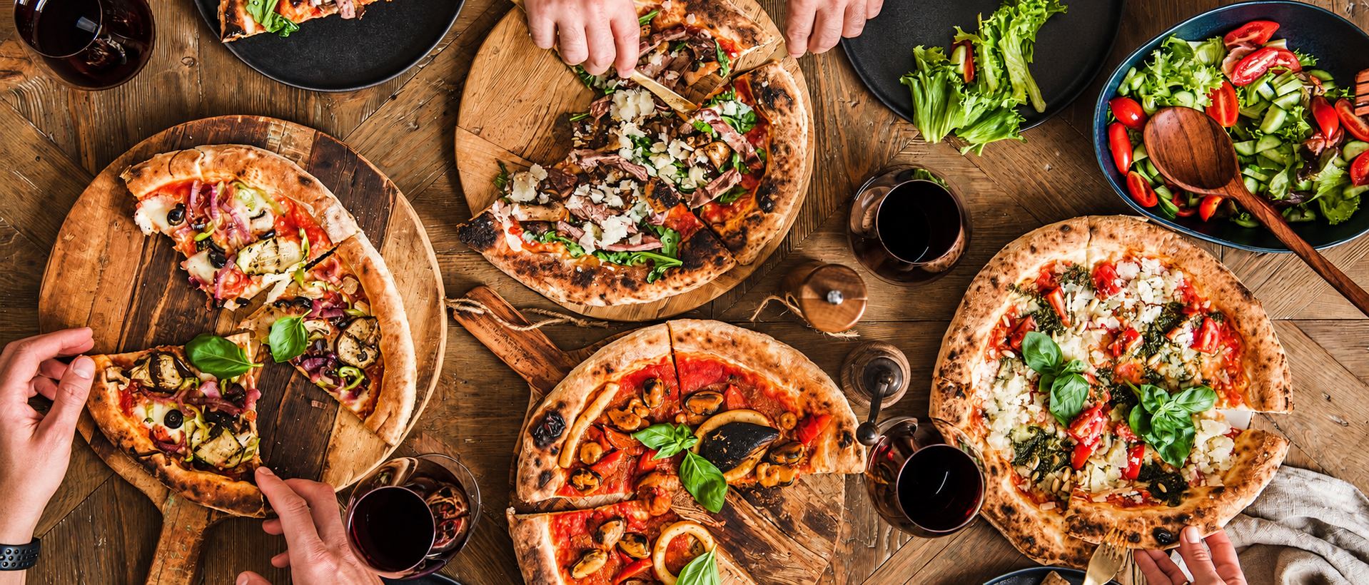 Table en bois couverte de pizzas, de salades et de verres, vue de dessus, des mains se servent